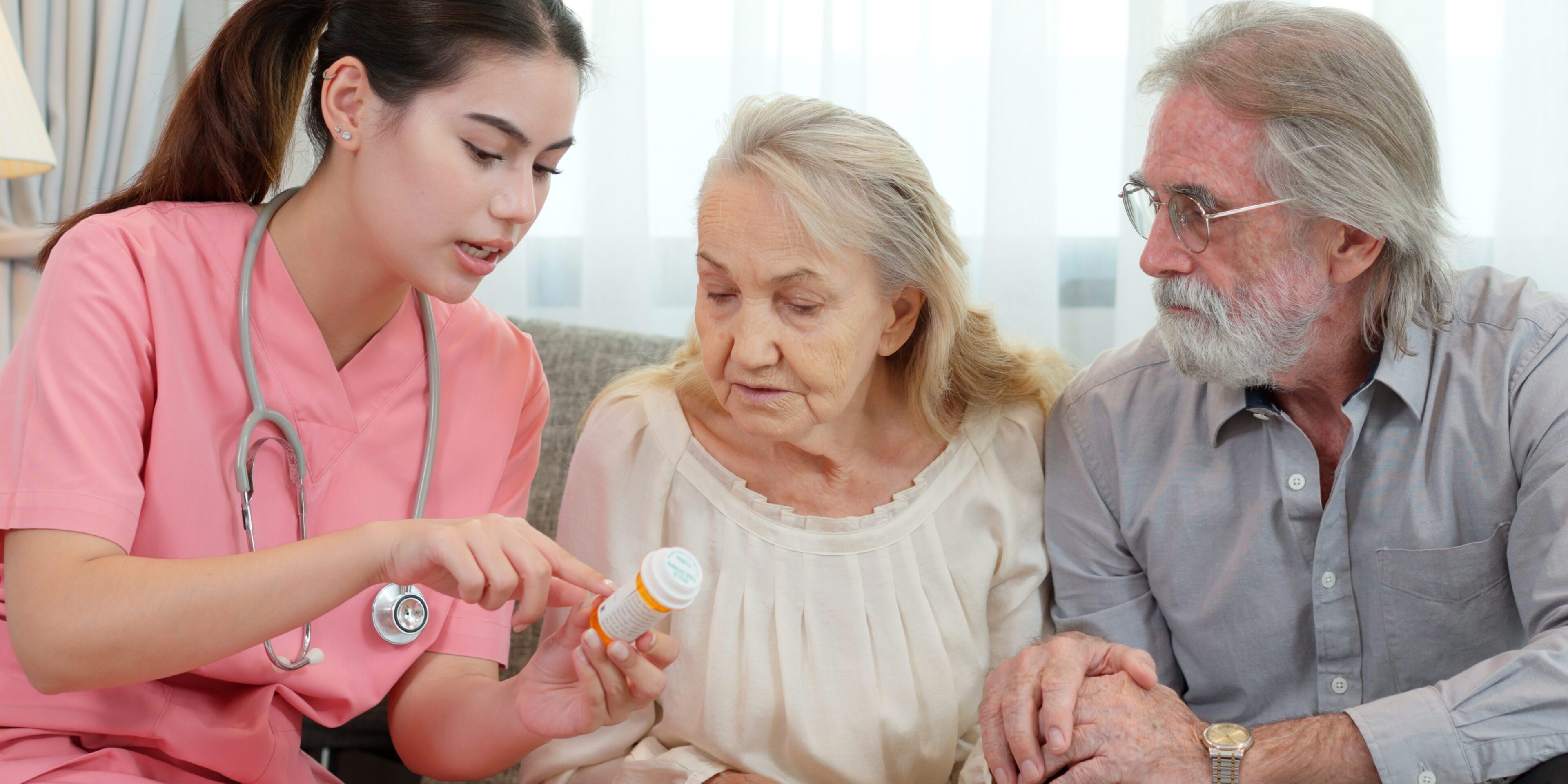 Caucasian young adult nurse is visiting elderly couple and holding a medicine bottle to explain how to use this medicine. Female caregiver is providing advice on health care to senior grandparents.