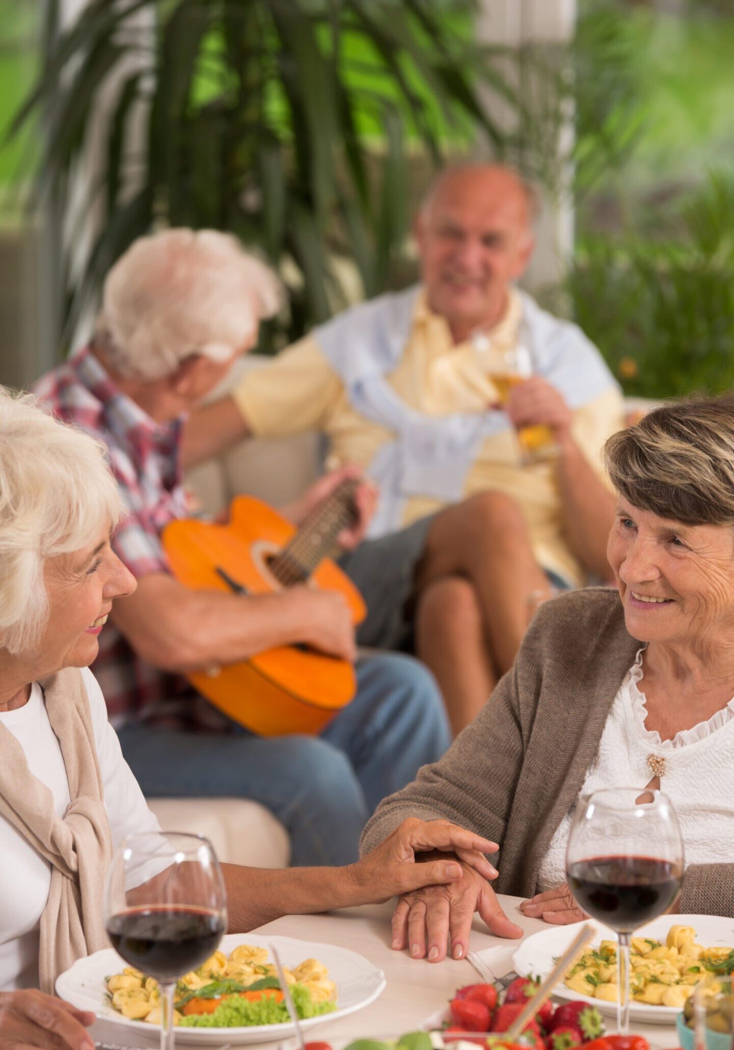 Two senior women, in the background elderly man playing guitar and his friend drinking wine